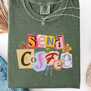 May include: Olive green t-shirt with the words "Send Coffee" in a colorful, collage-style design. The shirt has a crew neck and short sleeves. A silver necklace with a sun pendant is draped across the shirt.