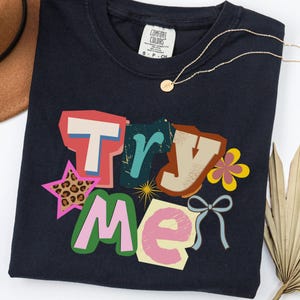 May include: Navy blue t-shirt with the words "Try Me" in a colorful, collage-style design. The design includes a star with a leopard print, a flower, and a bow. A gold necklace with a pendant is draped on the shirt.