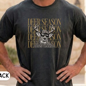 May include: A black short-sleeve t-shirt featuring a graphic of a deer wearing a baseball cap and sunglasses. The text DEER SEASON is printed in gold lettering four times in a stacked vertical pattern behind the deer illustration.