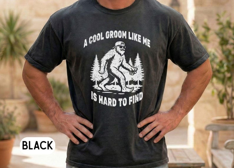 Op de afbeelding: Zwart t-shirt met een witte afbeelding van een Bigfoot-achtig wezen met een zonnebril en een telefoon. De tekst luidt: "A COOL GROOM LIKE ME IS HARD TO FIND".