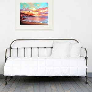 May include: A white metal daybed with a white mattress and a framed painting of a sunset over the ocean. The painting is in a white frame and is hanging on a white wall.