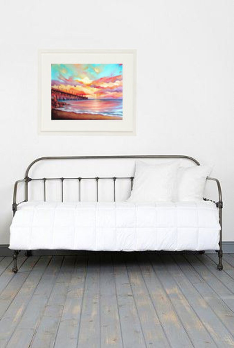 May include: A white metal daybed with a white mattress and a framed painting of a sunset over the ocean. The painting is in a white frame and is hanging on a white wall.