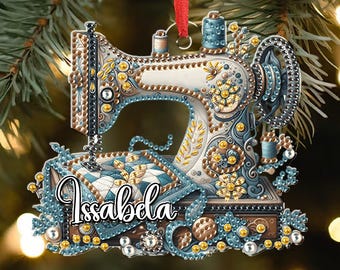 May include: A decorative Christmas ornament shaped like a vintage sewing machine. The ornament features intricate details in blue, gold, and white, with the name "Issabela" in cursive. It hangs from a red ribbon against a blurred background.