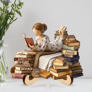 May include: A wooden figurine of a person reading a book while reclining on a stack of books. The figurine is wearing a floral patterned outfit. The text "Just a girl who loves books" is visible on the base. The name "Emily" is on a book.