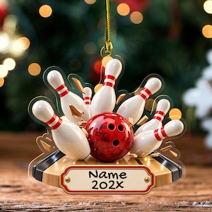 May include: A flat, hanging acrylic ornament featuring a red bowling ball striking white bowling pins with red stripes. The base is a wooden bowling lane section with a rectangular plaque displaying the text Name 202X in black font.