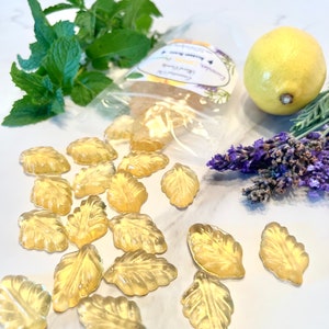 May include: A clear plastic bag filled with small, yellow, leaf-shaped candies. The candies are arranged in a pile on a white surface. There is a lemon, a sprig of lavender, and a sprig of mint in the background.