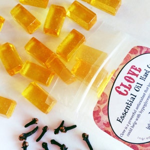 May include: A clear plastic bag filled with small, rectangular, yellow-amber colored hard candies. The bag has a label that reads "CLOVE Essential Oil Hard Candy". The text on the label says "Clove is a powerful antioxidant that has been shown to help with hypoglycemia". There are also several whole cloves scattered on a white surface below the bag.