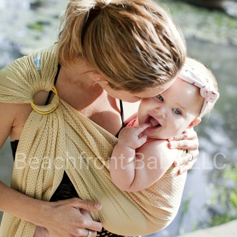 Puede incluir: Un portabeb&eacute;s beige con un anillo amarillo est&aacute; envuelto alrededor de una persona, que sostiene a un beb&eacute;. El beb&eacute; lleva un atuendo rosa y una diadema blanca. El beb&eacute; sonr&iacute;e y tiene el dedo en la boca. El portabeb&eacute;s tiene un dise&ntilde;o perforado.