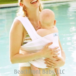May include: A woman in a white mesh baby carrier holds a baby in a swimming pool. The carrier has the text "Beachfront Baby LLC" on it. The woman is wearing a green and brown striped bikini bottom.