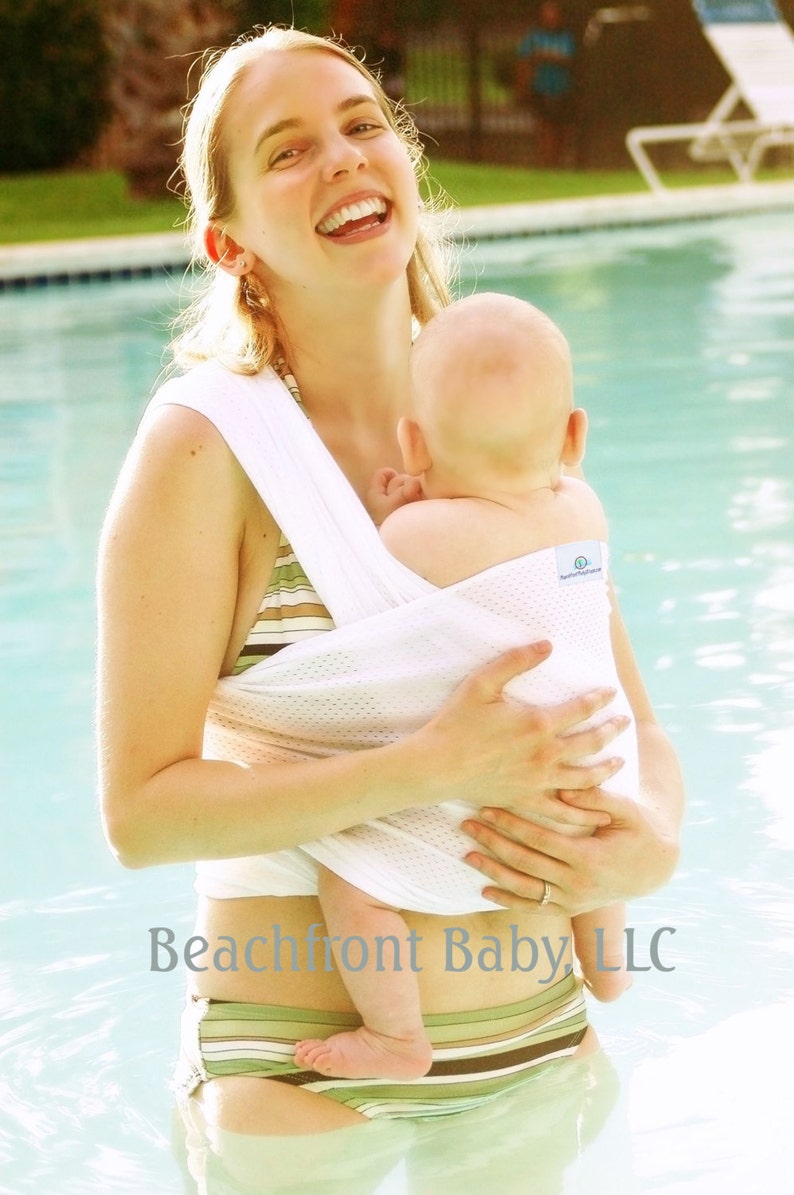 May include: A woman in a white mesh baby carrier holds a baby in a swimming pool. The carrier has the text "Beachfront Baby LLC" on it. The woman is wearing a green and brown striped bikini bottom.