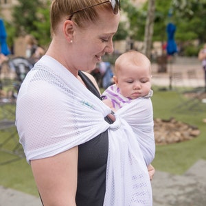 Puede incluir: Un portabeb&eacute;s de malla blanca con cierre de anillo plateado. El portabeb&eacute;s est&aacute; envuelto alrededor del usuario, sosteniendo a un beb&eacute; con una camisa a rayas. El fondo muestra un parque con &aacute;rboles y edificios.