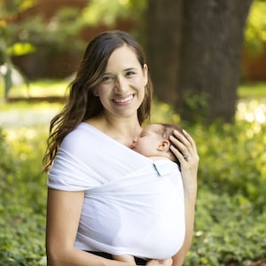 May include: A woman wearing a white stretchy wrap carrier with a baby sleeping inside. The woman is smiling and looking at the camera. The background is a blurred green and brown.