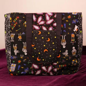 May include: A black tote bag featuring a patchwork design with Halloween-themed patterns including skeletons, mummies, spider webs, orange crescent moons, and yellow stars. The bag has two black fabric handles and a central pocket panel.