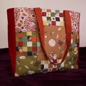 May include: A fabric tote bag featuring a patchwork design with Snoopy and Peanuts characters, autumn leaves, and pumpkins. The bag has orange handles, red side panels, and a central tan pocket with the text CHOMP CHOMP and PUMPKIN SPICE.