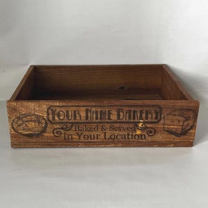 May include: A rectangular wooden bakery box with the text "YOUR NAME BAKERY Baked & Served in Your Location" etched on the front. The box is a warm brown color and features images of bread.
