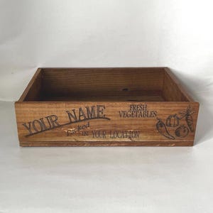 May include: A rectangular wooden crate with the words "YOUR NAME" and "FRESH VEGETABLES" printed on the front. The crate is brown and has a rustic appearance. The text "packed in YOUR LOCATION" is also visible.