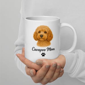 Könnte beinhalten: Weiße Keramiktasse mit einer detaillierten Illustration eines Cavapoo-Hundes mit flauschigem, goldbraunem Fell. Die Tasse trägt den Text "Cavapoo Mom" und einen Pfotenabdruck. Die Tasse hat einen großen Henkel.