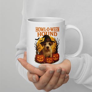 Può includere: Tazza in ceramica bianca con un design a tema Halloween. Il design include un cucciolo con un cappello da strega, zucche, pipistrelli e il testo "HOWL-O-WEEN HOUND" in arancione. La tazza è tenuta da una persona.