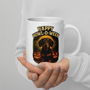 Può includere: Tazza in ceramica bianca con un design a tema Halloween. Presenta un bassotto nero e marrone, due zucche intagliate e la scritta "HAPPY HOWL-O-WEEN". Lo sfondo include una luna piena e pipistrelli.