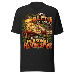May include: A black t-shirt featuring a graphic of a ball python resting on a red velvet cushion under a lamp. The text reads: MY BALL PYTHON ISN'T SPOILED. HE JUST HAS A PERSONAL HEATING STAFF. A small sign nearby displays 88 F.