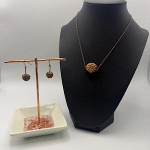 Puede incluir: Un conjunto de joyas con un collar y pendientes con un colgante esférico texturizado. El collar se exhibe en un maniquí negro, mientras que los pendientes cuelgan de un pequeño soporte. También hay un plato cuadrado blanco con detalles en rosa y naranja.