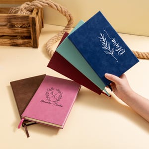 May include: A collection of personalized journals in various colors, including blue, burgundy, and pink. The journals feature embossed designs and text, with one displaying the name "Olivia" and another "Alexander + Claudia". Each journal has a ribbon bookmark.