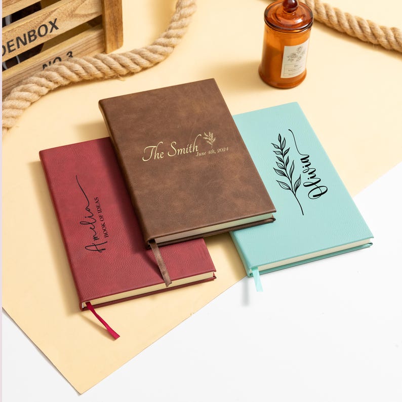 May include: Three personalized notebooks in burgundy, brown, and teal. The brown notebook is embossed with "The Smith" and a date. The burgundy notebook says "Amelia Book of Ideas". The teal notebook says "Olivia". Each has a ribbon bookmark.