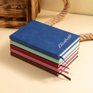 May include: A stack of five colorful notebooks. The top notebook is blue with the name "Elizabeth" in white script. The other notebooks are green, red, pink, and brown. Each notebook has a ribbon bookmark.