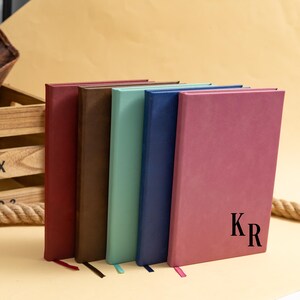 May include: A collection of six hardcover journals in various colors: burgundy, brown, seafoam green, navy blue, and mauve. The mauve journal has the initials "KR" printed on the cover. Each journal has a matching ribbon bookmark.
