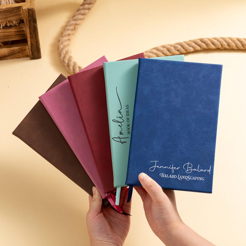 May include: A collection of personalized notebooks in various colors, including brown, pink, burgundy, and blue. The notebooks have text that reads "Amelia BOOK OF IDEAS" and "Jennifer Balard BALARD LANDSCAPING".