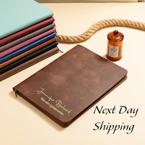 May include: A brown leather portfolio with a zipper closure, personalized with the name "Jennifer Balard" and "BALARD LANDSCAPING" in gold lettering. A stack of colorful notebooks and a decorative candle are also visible.
