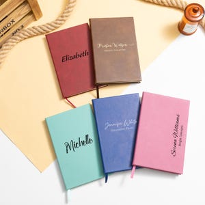 May include: Five personalized notebooks in various colors: red, brown, blue, pink, and teal. Each notebook features a name or text in a contrasting color. The notebooks have a ribbon bookmark.