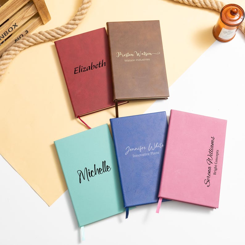 May include: Five personalized notebooks in various colors: red, brown, blue, pink, and teal. Each notebook features a name or text in a contrasting color. The notebooks have a ribbon bookmark.