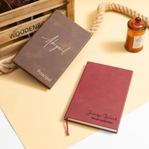 May include: Two personalized leather journals. One is brown with "Abigail" and "Principal" in gold lettering. The other is burgundy with "Jennifer Balard" and "BALARD LANDSCAPING" in black lettering. A candle and rope are in the background.