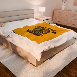 May include: A mustard yellow blanket with a black graphic design featuring a bird-like creature and the word "POWER". The blanket is folded on a bed with white sheets. The bed is in a well-lit room.