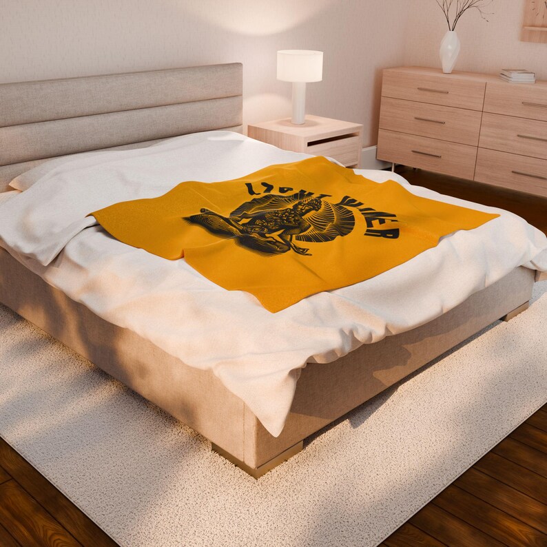 May include: A mustard yellow blanket with a black graphic design featuring a bird-like creature and the word "POWER". The blanket is folded on a bed with white sheets. The bed is in a well-lit room.