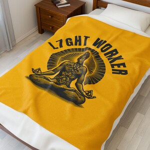 May include: Mustard yellow blanket with a black graphic of a meditating figure and the words "LIGHT WORKER". The design features a sunburst behind the figure. The blanket is folded over a white bed.