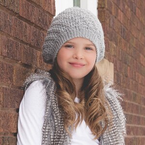 May include: A young person wearing a gray crocheted beanie and a white shirt with a gray and white faux fur vest.