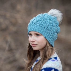 May include: A young person wearing a blue crocheted hat with a white pom pom. The hat has a textured design and is worn over long brown hair. The person is wearing a grey and blue patterned sweater.