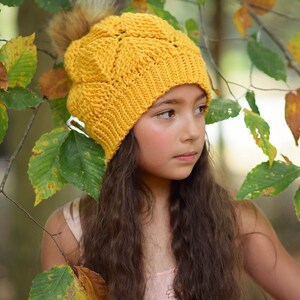 May include: A young girl wearing a yellow crocheted beanie with a fluffy pom pom. The beanie has a textured pattern and is worn over long brown hair. The girl is standing in a forest setting with green and yellow leaves.