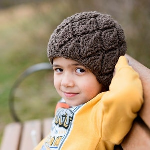 May include: A young person wearing a brown crocheted hat sits on a wooden bench. They are wearing a yellow sweatshirt with a graphic design on the front and grey pants. The person is looking at the camera and smiling.