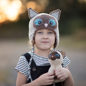 May include: A crocheted beanie hat designed to look like a Siamese cat with pointed ears, a dark brown face, and large blue eyes, featuring braided side tassels. The hat is paired with a matching small plush Siamese cat toy.