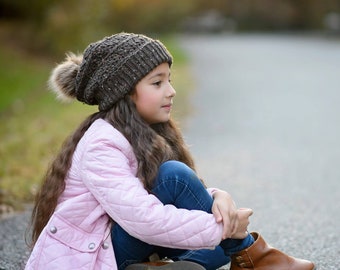 May include: A young girl wearing a brown knit beanie with a pom pom sits on a paved road. She is wearing a pink quilted jacket and blue jeans. The girl is looking off to the side.