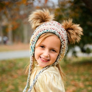 May include: A young person wearing a colorful knitted hat with two large brown fur pom-poms. The hat has ear flaps and braided ties. The person is also wearing a gold pleated dress.