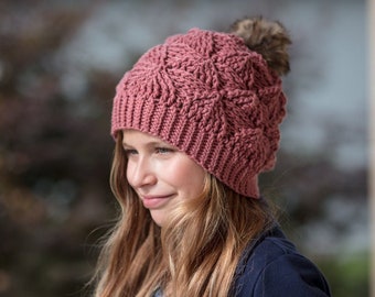 May include: A pink crocheted beanie with a white pom pom. The beanie has a cable knit pattern and is worn by a person with long blonde hair.