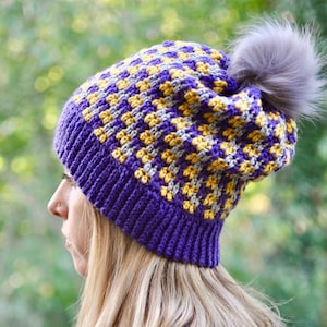 May include: A handmade purple, yellow, and gray knit beanie with a faux fur pom-pom. The hat features a ribbed brim and a textured pattern on the crown. The beanie is designed for warmth and style.
