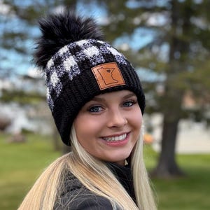 May include: A black and white checkered knit beanie with a faux fur pom-pom. The hat features a brown leather patch with a state outline. The beanie is worn by a person with long blonde hair.