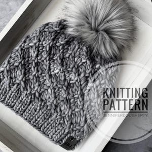 May include: A chunky knit beanie in a variegated grey yarn featuring a large, fluffy faux fur pom-pom on top and a small black rectangular tag labeled KBJ at the brim. The image includes a circular watermark that reads KNITTING PATTERN JENNIFERDOUGHERTY.