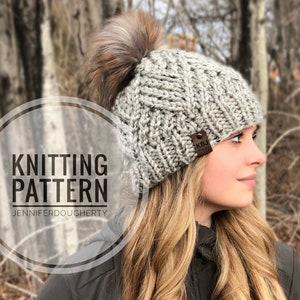 Könnte beinhalten: Graue Strickmütze mit braunem Kunstfell-Bommel. Die Mütze hat ein strukturiertes Muster und ein kleines braunes Lederetikett. Der Text "KNITTING PATTERN" und "JENNIFERDOUGHERTY" ist auf dem Bild zu sehen.