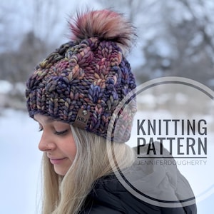 May include: A hand-knitted beanie in a mix of purple, pink, and yellow hues, topped with a fluffy, multi-colored pom-pom. The hat features a textured knit pattern and a small leather tag. Text on the image reads "Knitting Pattern" and "JenniferDougherty".